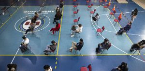 People in a gymnasium sitting in socially distanced chairs