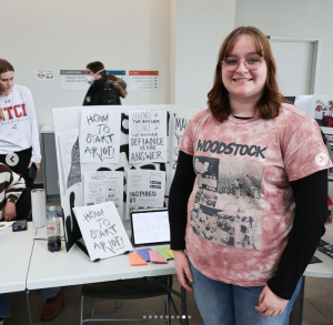 Student wearing a Woodstock tee standing next to a poster board that says "How to start a riot."